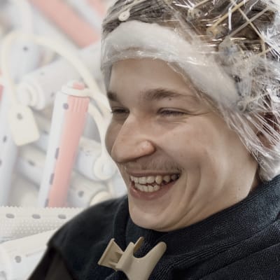 Samuli Souru nauraa. Hänellä on päässään permanenttirullat, jotka on kääritty muoviin. Kuvan taustalle on lisätty permanettirullia.