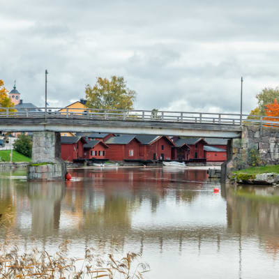 En bro går över en spegelblank å, i bakgrunden skymtar röda bodar