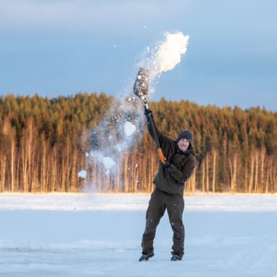 Iloinen Paul Lancaster heittää lumilapiolla lunta ilmaan päänsä yläpuolelle Saimaan jäällä. 
