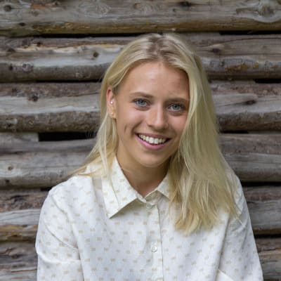 En ung kvinna med blont hår och vit skjorta sitter framför en lada gjord av stockar. Det är sommar.