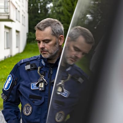 Poliisi Björn Borg seisoo poliisiautonsa vieressä ja hänen kuvansa heijastuu auton ikkunasta.