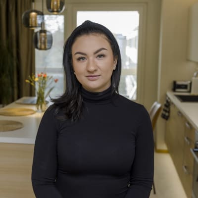 Nuori nainen mustissa vaatteissa seisoo kodin keittiössä ja katsoo suoraan kameraan.