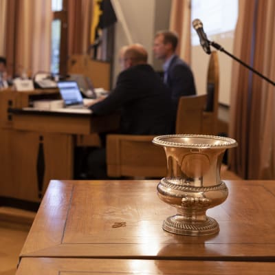 Mikkelin kaupunginjohtajan valinnassa käytetty kiiltävä metalliastia seisoo puupöydällä Mikkelin kaupunginvaltuuston salissa. Taustalla siintää valtuutettuja.