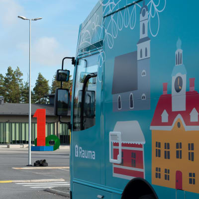 Värikäs kirjastoauto, jonka kyljessä on kuva raatihuoneesta. Taustalla koulurakennus, jonka seinässä lukee Pohjoiskehän koulu.