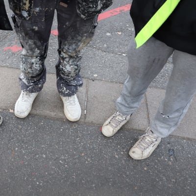 Kolmen nuoren jalat asfaltilla. Kaikilla on nauhakengät jalassaan. Keskimmäisellä on maalin tahrimat housut. 