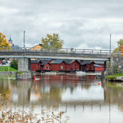 En bro går över en spegelblank å, i bakgrunden skymtar röda bodar