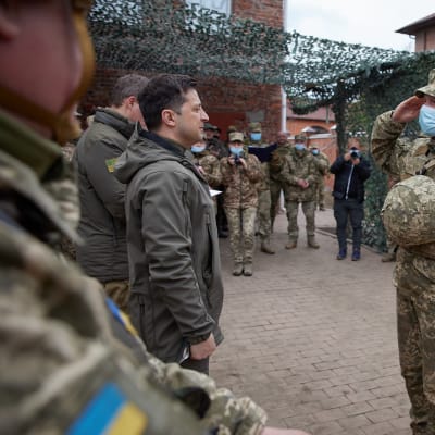 Ukrainas president Volodomyr Zelenskyj står tillsammans med en grupp soldater framför honom står en soldat i ansiktmask som gör honnör.