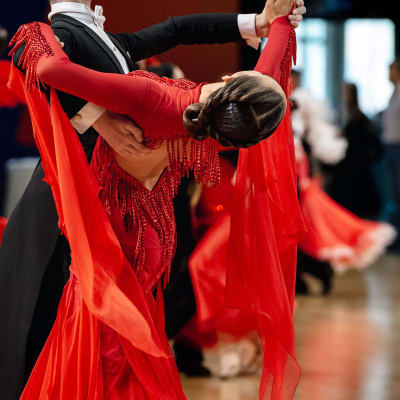 Ett par som dansar rumba. Följaren är iklädd en röd klänning, med håret stramt uppsatt i en knut, och böjer med ansiktet bort från kameran. Ledaren är iklädd kostym.