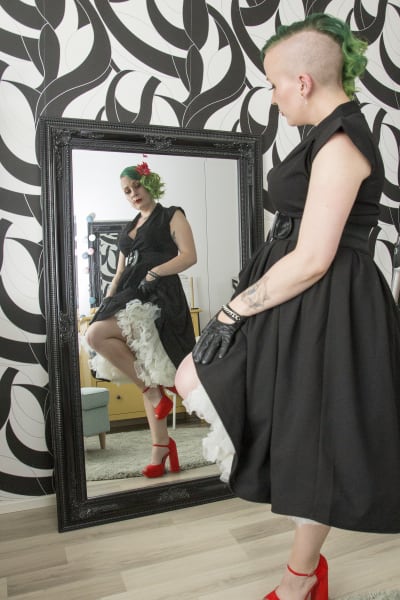 En kvinna med grönt hår står framför en helkroppsspegel och speglar sig.
