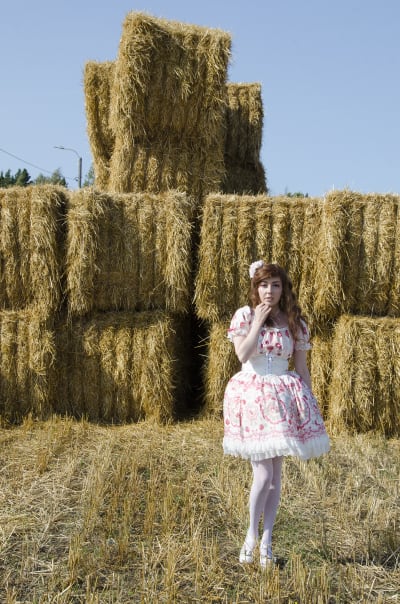 En kvinna klädd i så kallade Lolitakläder - en fluffig vit och rosa klänning med spetsar på. Hon har vita högklackade skor och blommor i håret. Hon står framför en massa höbalar.