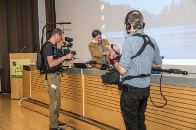 Kuvassa yksi mies äänittää ja toinen kuvaa kun ufotutkija Heikki Kulju valmistelee puhuja Gosiaa ufoseminaarin alkamiseen.