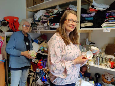 Två kvinnor står i ett rum som används som loppmarknad. Det är massor av saker på hyllorna. De heter Taina Martikainen och Tanja Kakko.