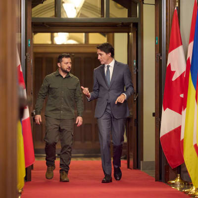 Zelenskyi ja Trudeau kävelevät vierekkäin käytävällä, jota reunustavat lippurivit.