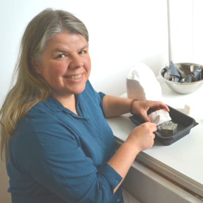Kvinna i blått sitter vid bord och skrapar med kniv mot sten. Hon ler mot kameran.