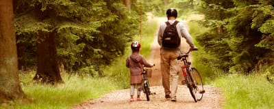 En vuxen och ett barn leder sina cyklar längs en väg i skogen.