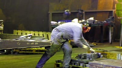 Nedre kroppen av person i overall lyfter metallföremål på fabriksgolv.