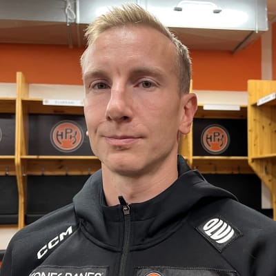 Mustaan huppariin pukeutunut Juuso Hietanen lähikuvassa jääkiekkojoukkueen pukukopissa, jonka seinällä oranssisia HPK-logoja.
