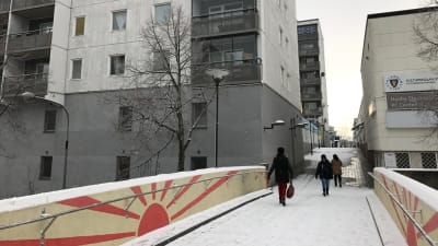Tre personer promenerar på en gångbro mellan höga bostadshus. Det är snö på marken.