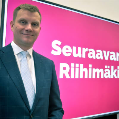 Mies hymyilee 7ison videonäytön edessä, jossa on suurella teksti Seuraavana Riihimäki.
