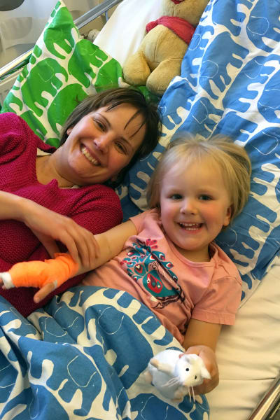 Mamma och hennes treåriga dotter ligger på en sjukhussäng, de ler mot kameran