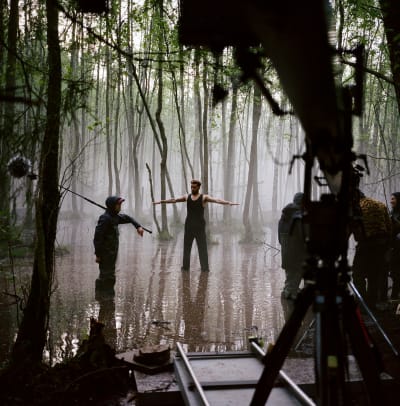 En man med händerna utsträcka står i en vattenpöl med en filmare framför sig.