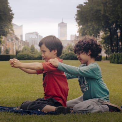 Två små pojkar, Aatos Amine, sitter bakom varandra på en gräsmatta i en park och leker tåg. Stillbild från dokumentären Aatos ja Amine.