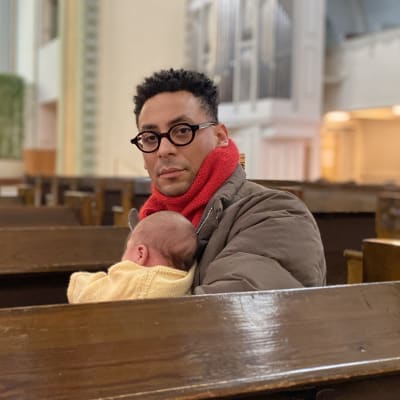 Sawandi Groskind sitter i en kyrkbänk iklädd en dunjacka och en halsduk med ett spädbarn i famnen.