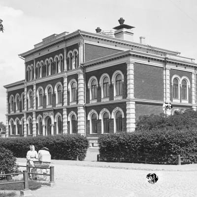 Mustavalkoinen kuva, jossa näyttävä tiilinen koulurakennus 1800-luvun lopulta pensasaitojen ympäröimänä, etualalla mukulakivinen risteys.