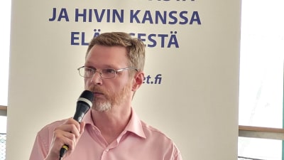 en blond och skäggig medeålders man sitter på en soffa och talar i mikrofon inför publik