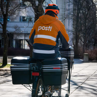 En brevbärare på cykel. Brevbäraren har på sig en orange rock med texten "Posti". Hen har ryggen vänd mot kameran.
