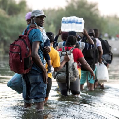 Människor går i vattnet i en flod mellan Mexiko och USA. En person håller ett paket med stora vattenflaskor på huvudet.