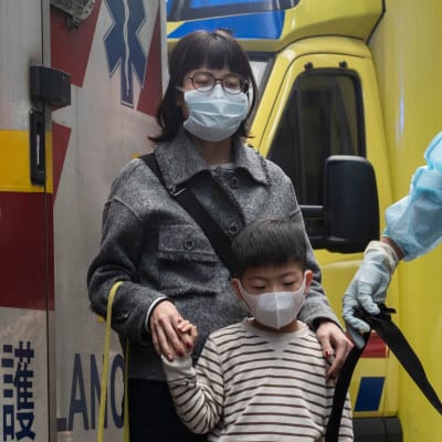 Sjukvårdare på gatan i blåa plastdräkter framför en ambulans. Bredvid står en kvinna och ett barn med ansiktsmasker.