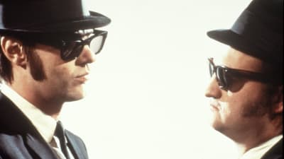 The Blues Brothers eli näyttelijät Dan Aykroyd ja John Belushi mustissa puvuissaan, mustissa laseissaan ja mustat hatut päässä sivukuvassa vastatusten valkoista taustaa vasten.
