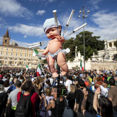 Bild på demonstranter på ett torg i rom som håller upp en docka med sprutor instuckna i armarna.