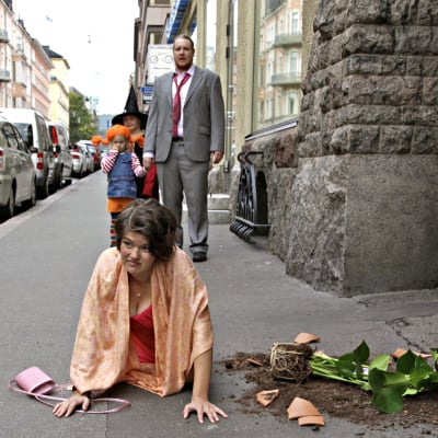Joanna Haartti kompastuneena kadulla rikkinäinen kukkaruukku vierellään, Santtu Karvonen ja lapset seisovat taustalla. Kuva lyhytelokuvasta Pitääkö mun kaikki hoitaa?.