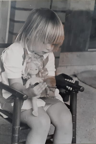 Mela, Mikaela Nyholm som barn med en docka i sin famn, sittande i en liten gungstol. Svartvit bild.
