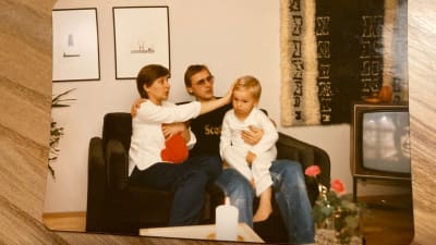 Perhe istuu pienellä mustalla sohvalla olohuoneessa, vauva äidin sylissä, ja pieni poika isän sylissä.