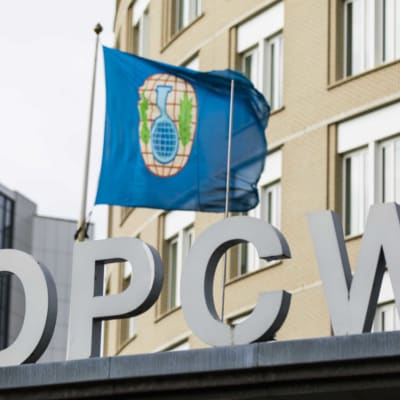 Kuvassa OPCW:n logo ja lippu, joka heiluu päämajan fasadissa.