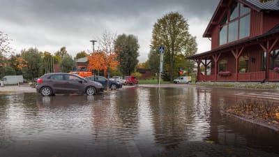 Bilar på en översvämmad parkeringsplats. Höstlandskap, en stor röd träbyggnad till höger. 