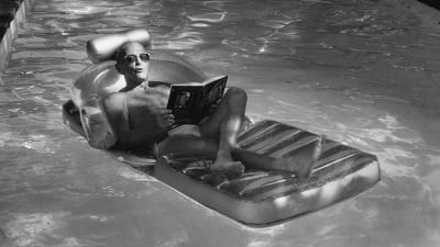 Svartvit bild av Schein i en pol där han på en badmadrass läser en bok.