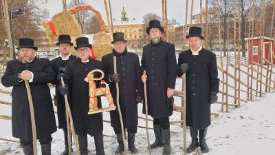 Sex svartklädda herrar i cylinderhattar står framför den jättelika Gävlebocken i Gävle.