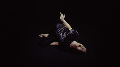 En dansare i svart t-skjorta ligger på ett svart golv.