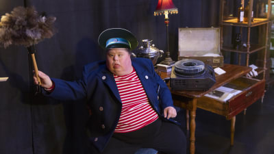 En skådespelare klädd som museivakt viftar med en dammvippa. 