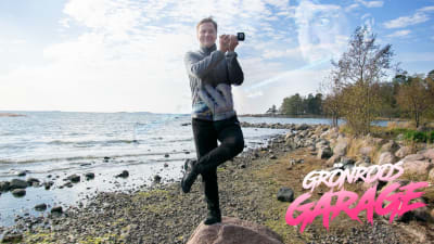 Carl-Kristian Rundman poserar på ett ben på strandsten och låtsas spela fläöjt med Yle-mikrofon. Ian Andersons ansikte bland molnen.