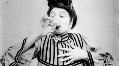 Nainen vanhanaikaisissa vaatteissa juo lasista puolilähikuvassa. Kuvakaappaus 1900-luvun alun elokuvasta Rouvan mielihalut.