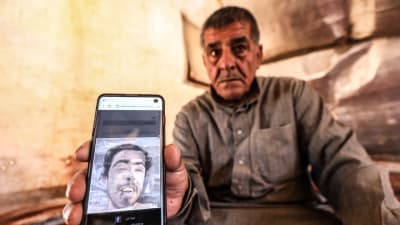 En sorgsen äldre man i gråbrun skjorta håller fram en mobil med en bild på en yngre man.