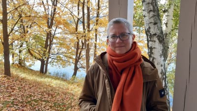 Litteraturforskaren Rita Paqvalén med stor orange halsduk i ett höstligt landskap med ett vattendrag i bakgrunden. 2021