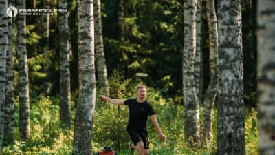En svartklädd man springer i skogen och ler. En frisbee flyger ovanför hans huvud. 