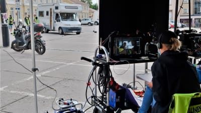 Bild av filmarbetare som ser på en skärm där skådespelarna syns. I bakgrunden finns en husbil där skåespelarna i verkligheten finns. 