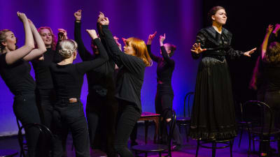 En grupp unga svartklädda kvinnor dansar på en teaterscen. Längst till höger står en kvinna i svart klänning på en stol. 